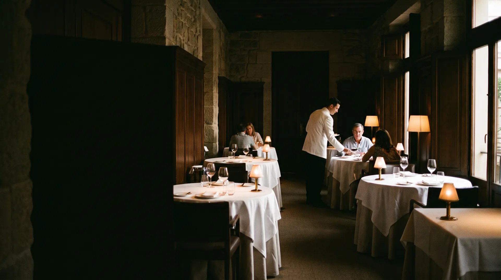 Comedor amplio de un restaurante elegante durante la cena, con mesas redondas de mantel blanco, lámparas de mesa y un camarero de chaqueta blanca sirviendo